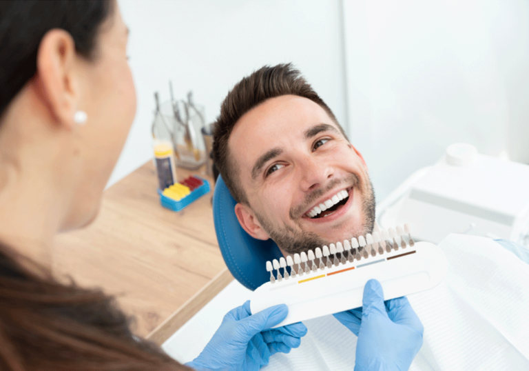 Close-up of a dentist in blue gloves holding a shade guide near a patient's teeth in a dental chair, with blurred dental tools and supplies visible in the background.