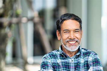 The image shows an older man wearing a checkered shirt in an outdoor setting. Trees and a building structure are faintly visible in the background.